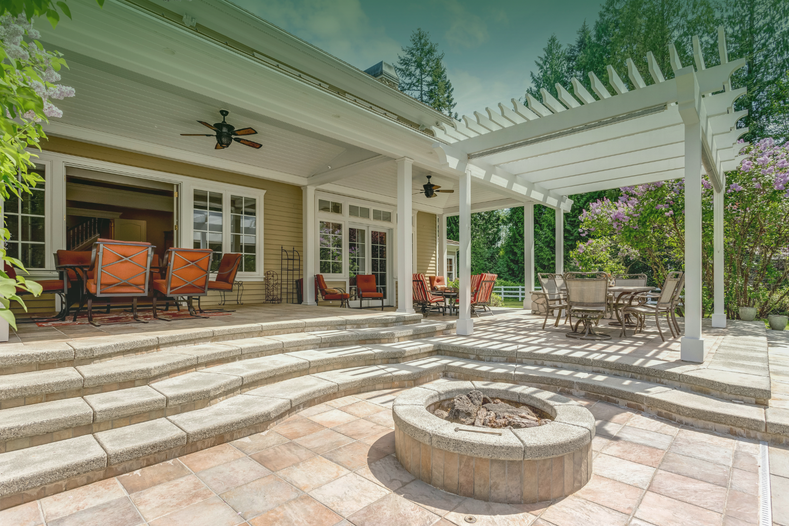 Backyard paver patio with white pergola, stone fire pit, and outdoor seating set into a forested yard.