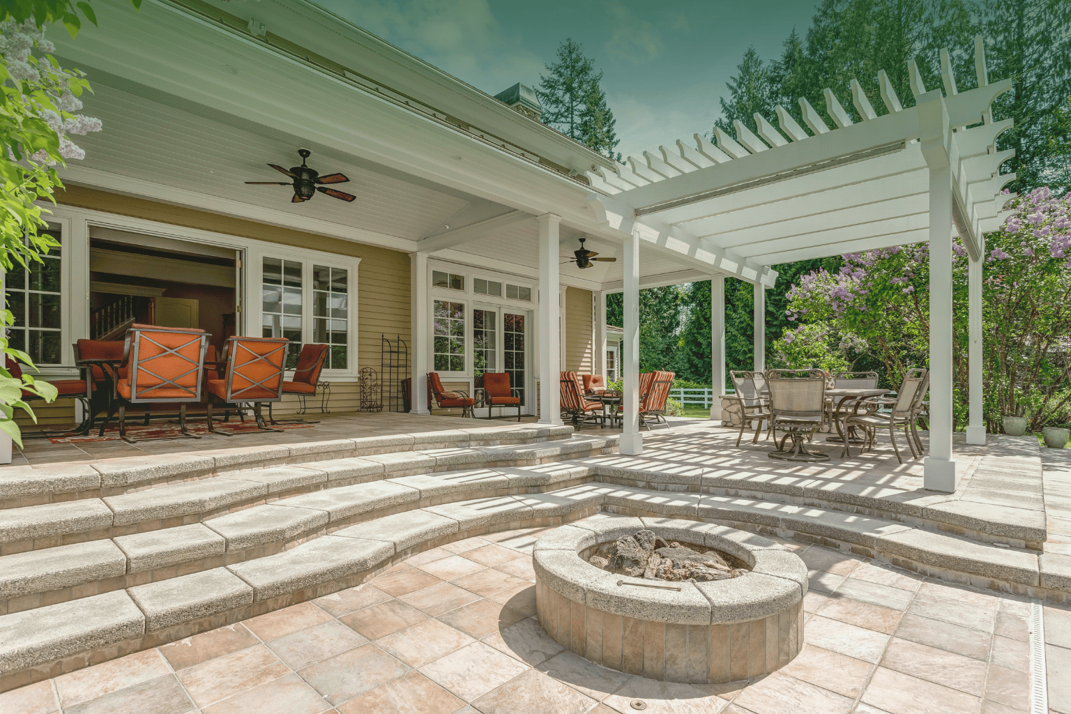Backyard paver patio with white pergola, stone fire pit, and outdoor seating set into a forested yard.