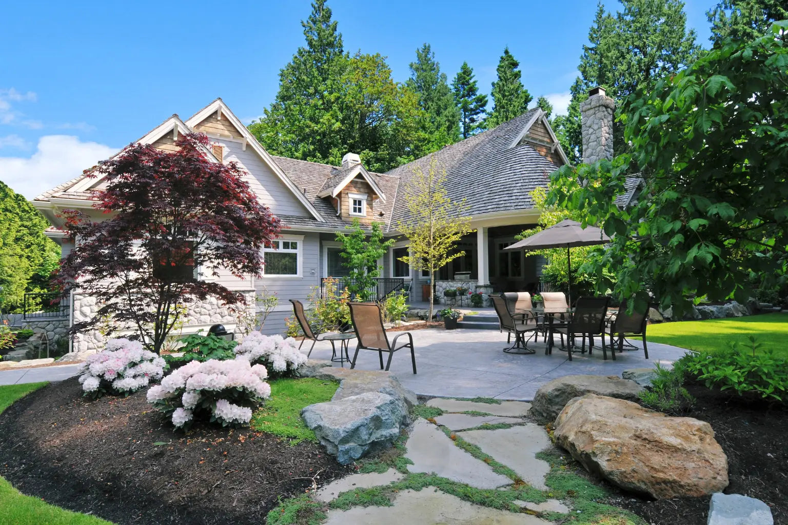 Two-story home with stone walkway, paver patio, manicured lawn, hydrangea borders, and rock landscaping.