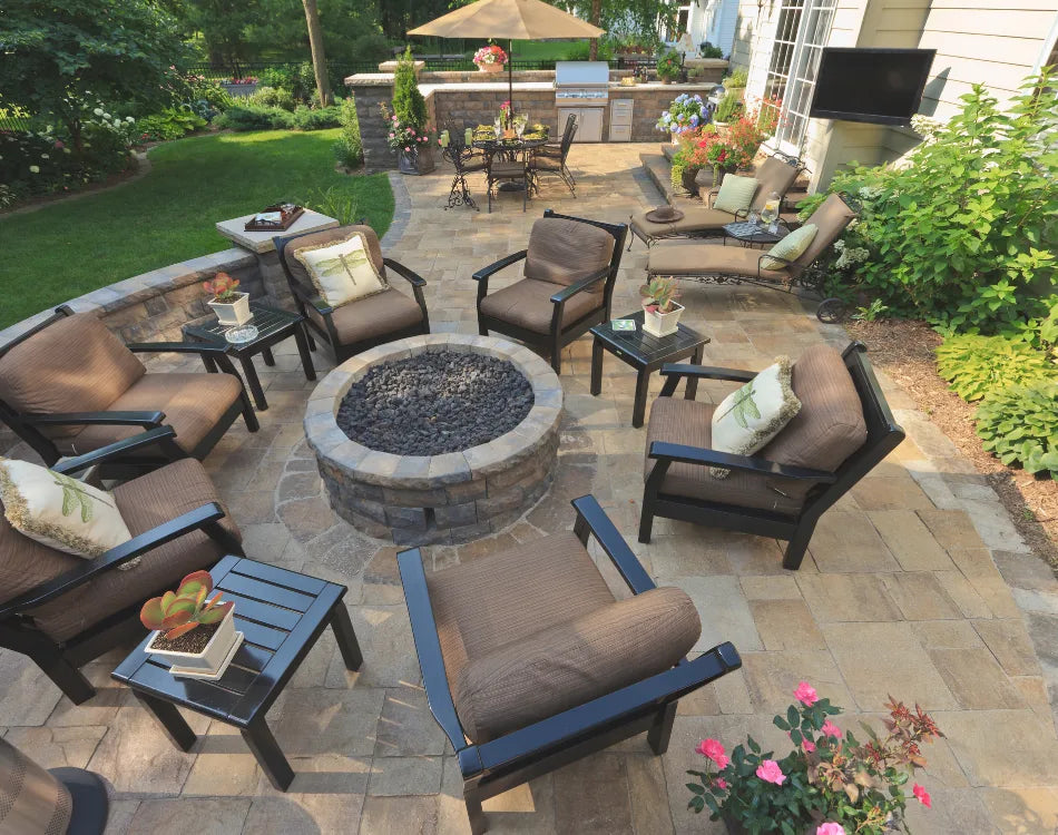 Spacious paver patio with circular fire pit, brown wicker armchairs, market umbrella, and lush garden borders.