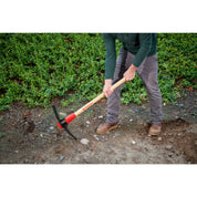 Gardener using pick mattock with two-handed swing to break up soil for a planting bed.
