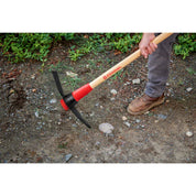 Gardener using pick mattock to dig at the edge of a paved walkway.