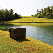 Dark brown aeration system unit by a pond, idyllic rural setting.
