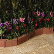 Red brick garden edging bordering vibrant pink, purple, and red flowers on a beige stone patio.
