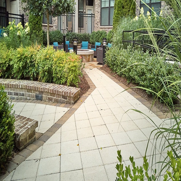 Off-white square pavers create a tranquil patio pathway, bordered by lush greenery and brick accents.
