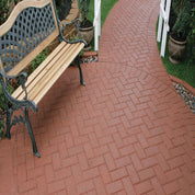 Red paver stones create a herringbone pattern walkway beside a wooden garden bench.
