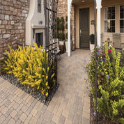 Tan pavers create a walkway leading to a home's entrance, bordered by vibrant yellow and multicolored shrubs.
