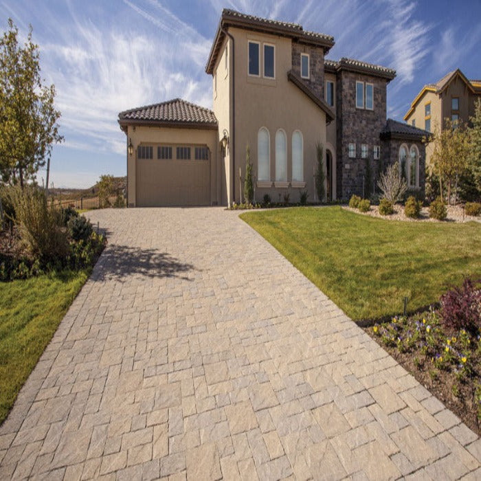 Tan paver stones create a driveway leading to a beige house with green lawn.
