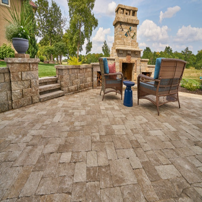 Outdoor patio scene with tan pavers, stone retaining wall, and wicker chairs around a stone fireplace.
