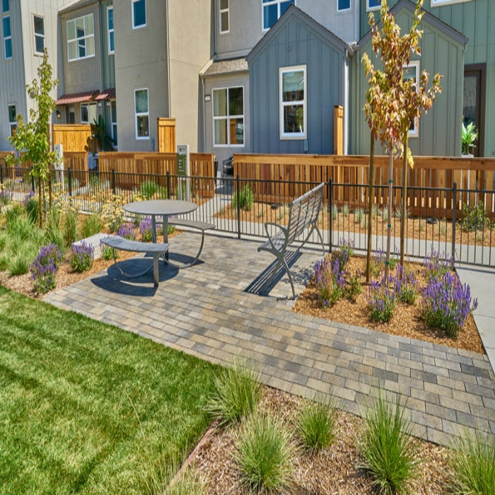 Outdoor patio scene with gray pavers, metal picnic table and bench, landscaping, and homes in background.
