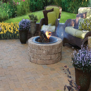 Stone fire pit burning on a patio, surrounded by wicker furniture and blooming plants.
