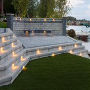 Evening view of a multi-level stone patio with integrated lighting, overlooking a lake.
