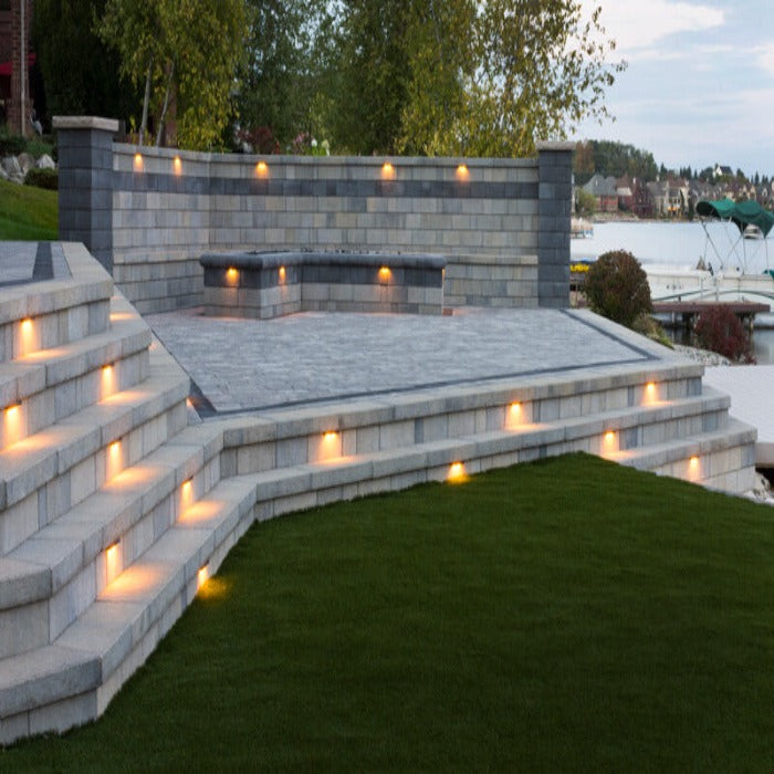 Evening view of a multi-level stone patio with integrated lighting, overlooking a lake.
