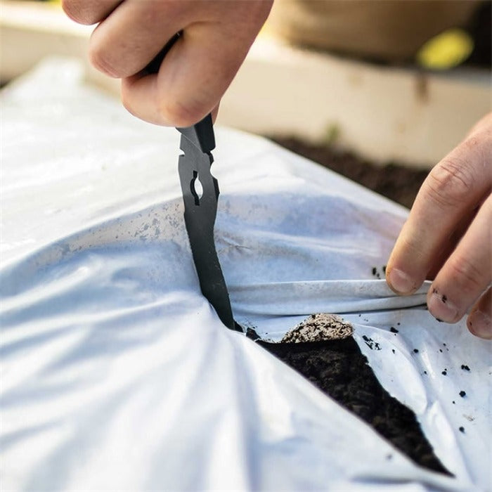 Hands using black gardening tool to cut a hole in white landscape fabric above dark soil.
