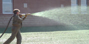 Person applying bonded fiber matrix to a lawn using a specialized sprayer near a brick building.
