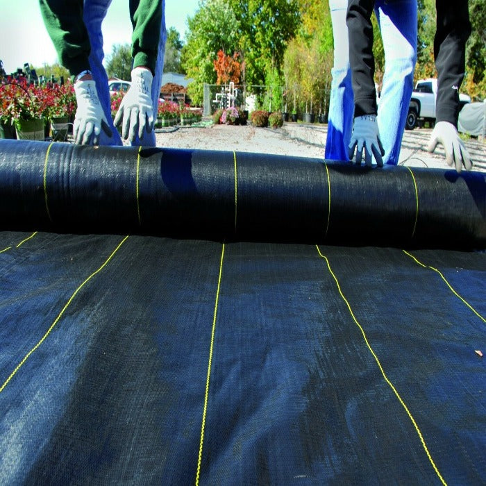 Black landscape fabric unrolling outdoors, showing reinforced yellow stitching.
